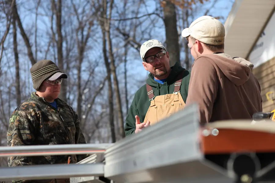 Two apprenticeship students and instructor in winter clothing discuss outdoors near construction materials.