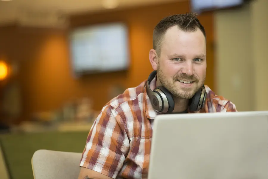 Male student smiling with headphones using a laptop in a blurred office setting.