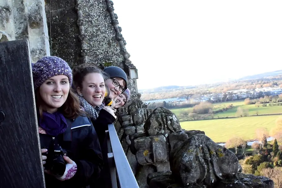 Three smiling students in London wearing winter clothes on a stone structure, scenic landscape in background.