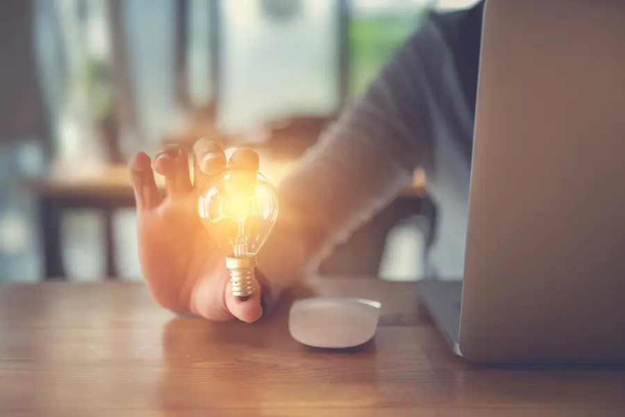 Hand holding a glowing light bulb near a laptop on a wooden desk.