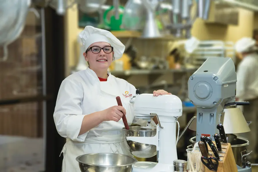 Student chef in white uniform using a stand mixer in a professional kitchen.
