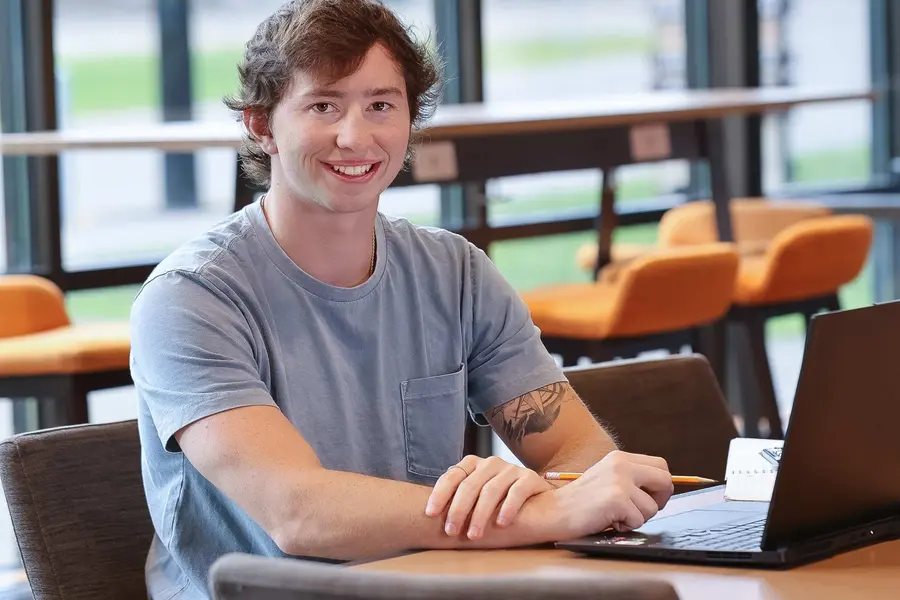 Scholarship Recipient Reuben Guzik smiling, seated at a table with a laptop in a bright lounge setting.