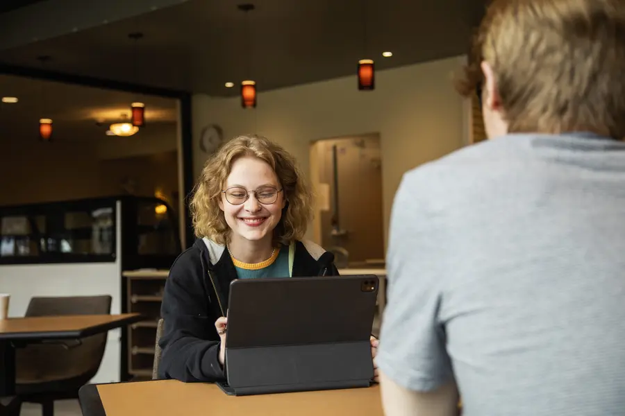 Two students talking at a table, one smiling with a tablet.
