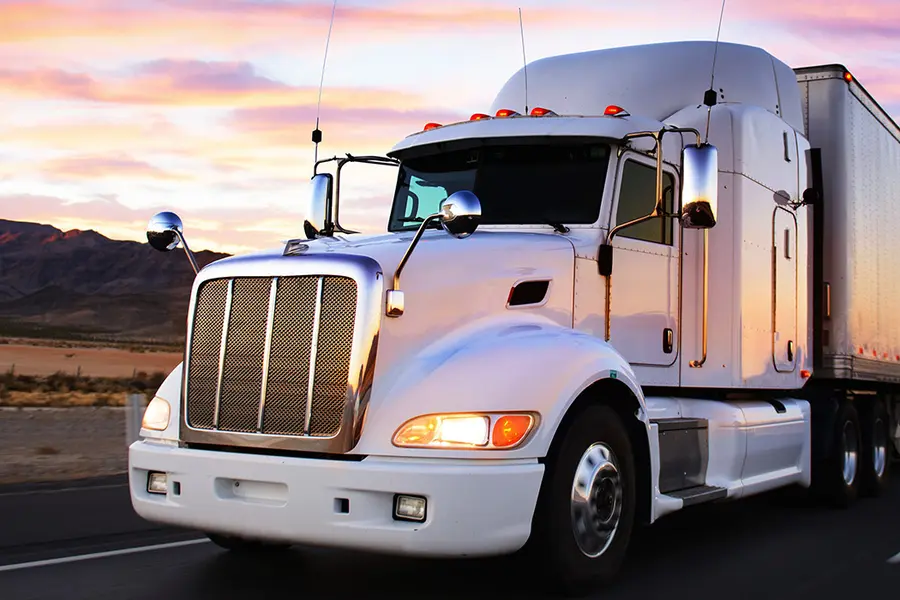 White semi-truck driving on a highway at sunset with mountains in the background.