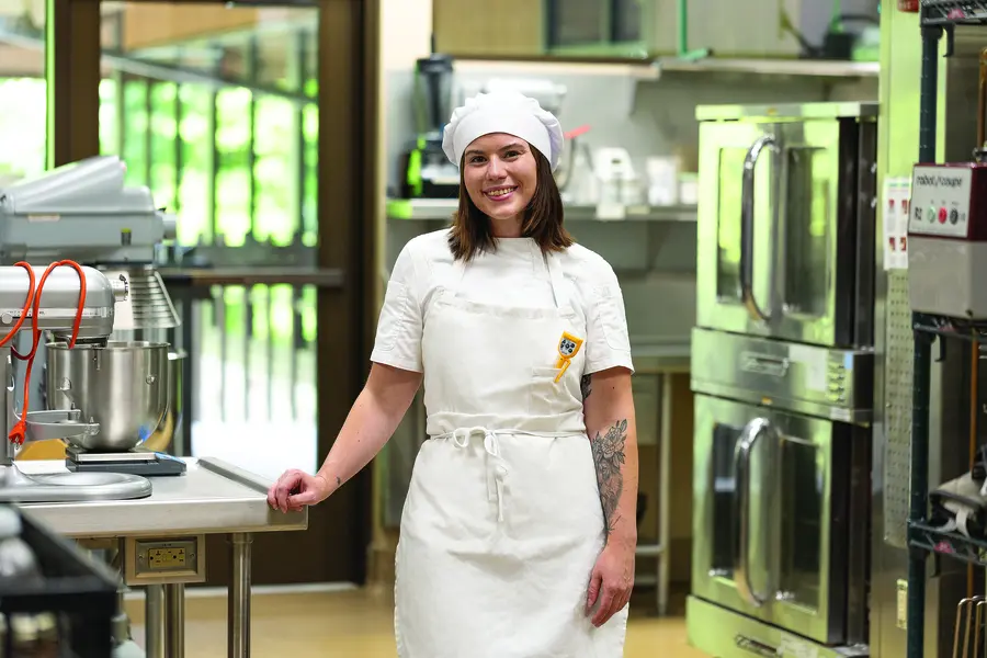 Culinary student Mykah Driscoll, smiling in a professional kitchen, wearing a white apron and hat.